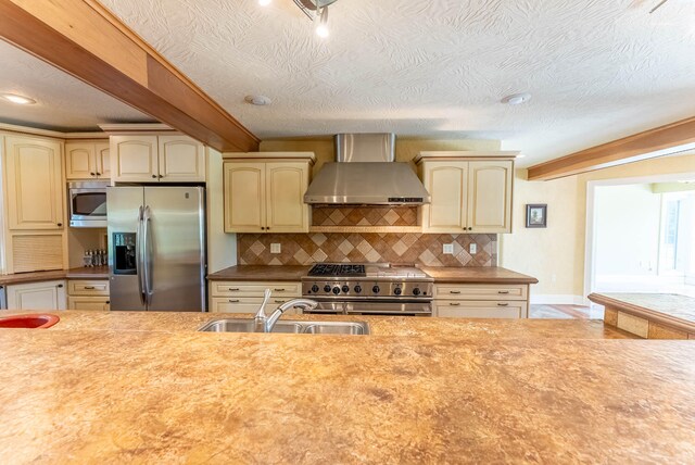 kitchen featuring cream cabinets, beamed ceiling, appliances with stainless steel finishes, and wall chimney range hood