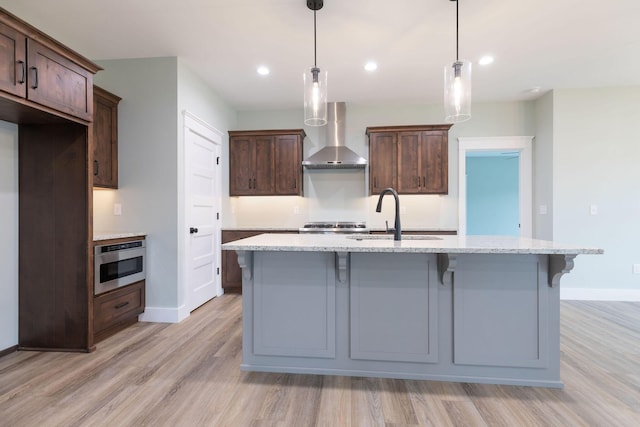 kitchen with an island with sink, pendant lighting, wall chimney exhaust hood, dark brown cabinetry, and sink