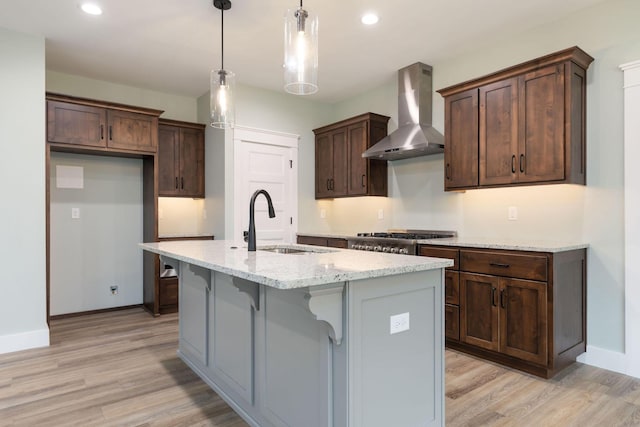 kitchen featuring a center island with sink, wall chimney range hood, decorative light fixtures, light stone countertops, and sink