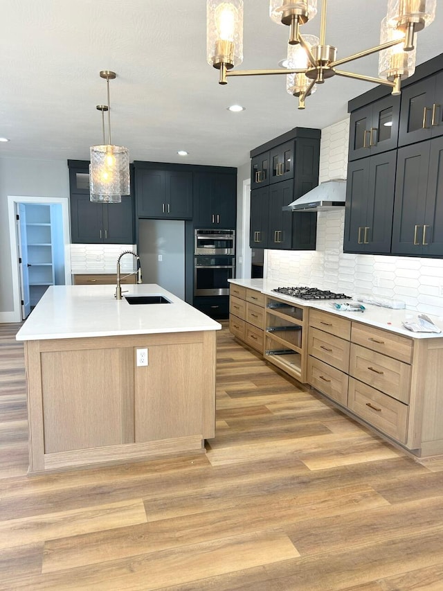 kitchen with decorative backsplash, appliances with stainless steel finishes, light brown cabinets, pendant lighting, and an island with sink