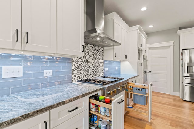 kitchen featuring wall chimney range hood, light hardwood / wood-style floors, light stone counters, appliances with stainless steel finishes, and white cabinetry