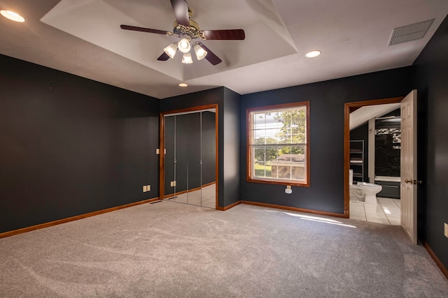 unfurnished bedroom with ceiling fan, light colored carpet, a raised ceiling, a closet, and connected bathroom