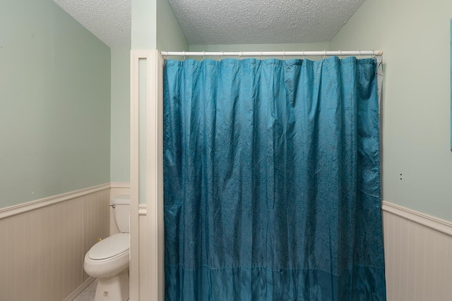 full bath with a shower with curtain, a textured ceiling, a wainscoted wall, and toilet