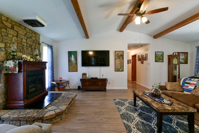 living room featuring ceiling fan, vaulted ceiling with beams, hardwood / wood-style floors, and a stone fireplace