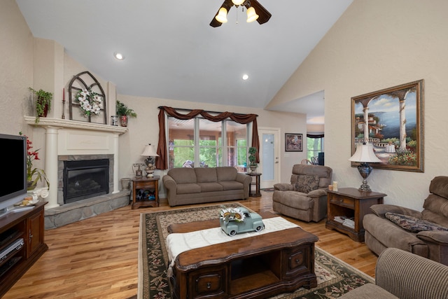 living room with ceiling fan, a stone fireplace, light hardwood / wood-style flooring, and high vaulted ceiling