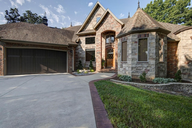french provincial home with stone siding, an attached garage, a shingled roof, and concrete driveway
