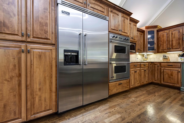 kitchen featuring dark wood-style flooring, decorative backsplash, glass insert cabinets, light stone countertops, and appliances with stainless steel finishes