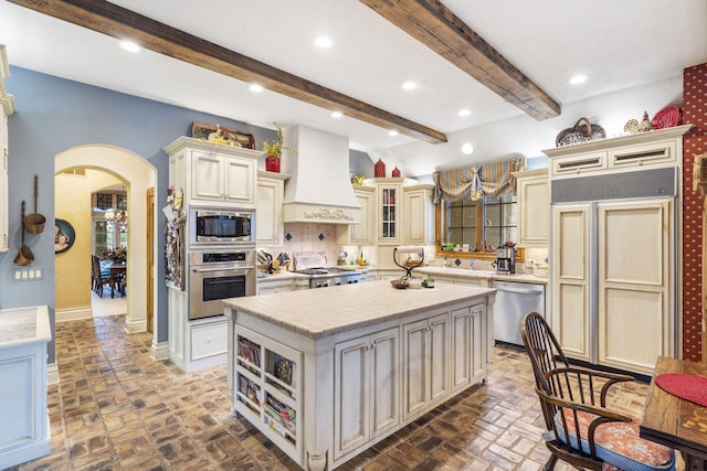 kitchen featuring a kitchen island, custom range hood, built in appliances, beam ceiling, and cream cabinetry