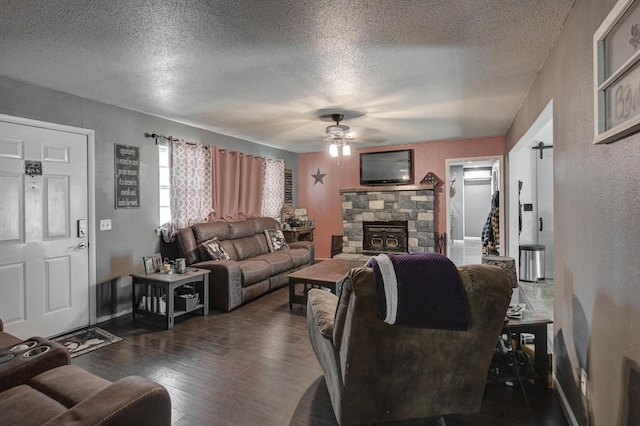 living room featuring a ceiling fan, a stone fireplace, a textured ceiling, a textured wall, and dark wood-type flooring