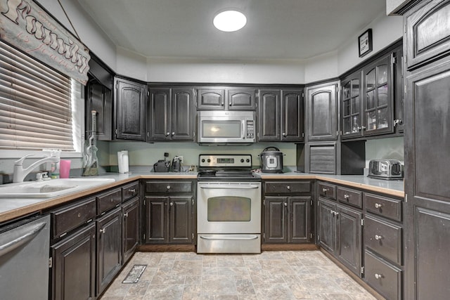 kitchen with a sink, glass insert cabinets, light countertops, stone finish flooring, and appliances with stainless steel finishes