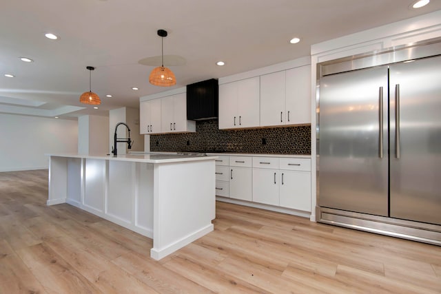 kitchen featuring light wood-type flooring, an island with sink, built in fridge, white cabinetry, and decorative light fixtures