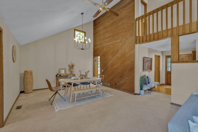 dining space featuring ceiling fan with notable chandelier, light colored carpet, high vaulted ceiling, and wood walls