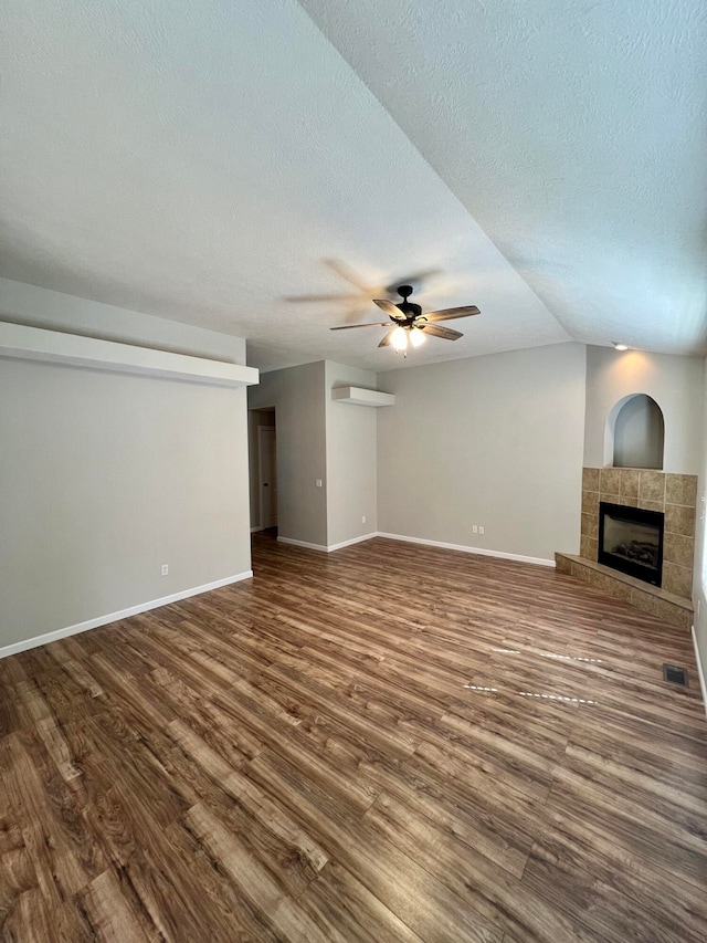 unfurnished living room featuring ceiling fan, wood-type flooring, a tiled fireplace, vaulted ceiling, and a textured ceiling