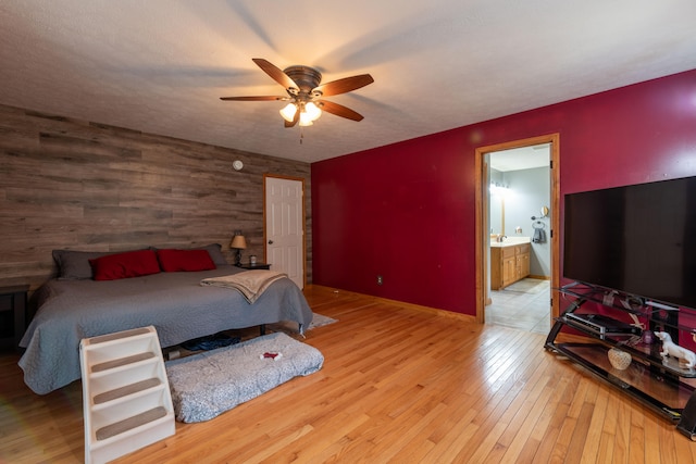 bedroom with ensuite bath, ceiling fan, wooden walls, and light hardwood / wood-style flooring