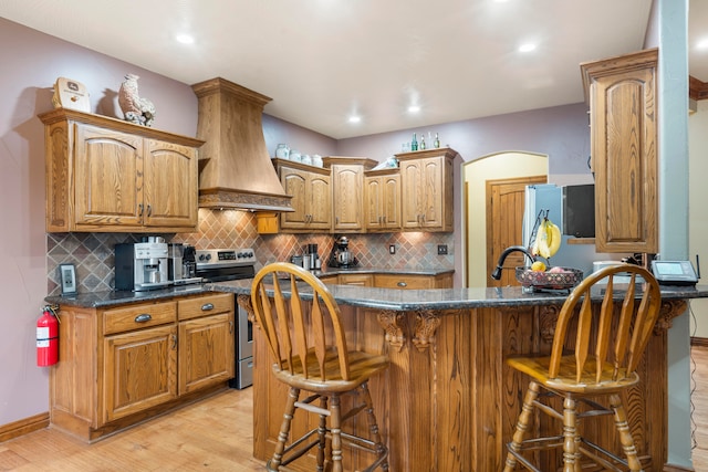 kitchen with stainless steel range with electric cooktop, light hardwood / wood-style flooring, a kitchen breakfast bar, backsplash, and premium range hood