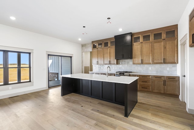 kitchen with light countertops, backsplash, brown cabinetry, an island with sink, and stainless steel gas range oven