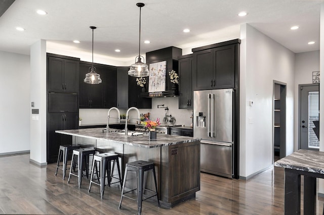 kitchen with pendant lighting, an island with sink, high end fridge, wall chimney exhaust hood, and dark wood-type flooring