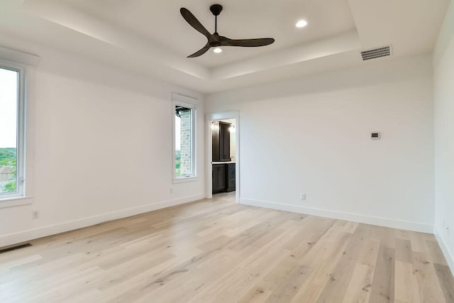 empty room with light wood-type flooring, a healthy amount of sunlight, ceiling fan, and a raised ceiling