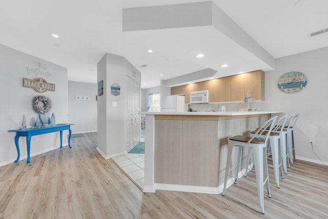 kitchen featuring light wood-type flooring, white appliances, light brown cabinetry, a breakfast bar area, and kitchen peninsula