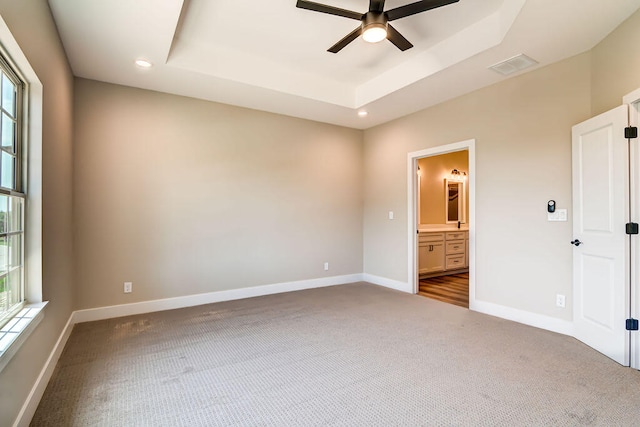 unfurnished bedroom featuring ceiling fan, a tray ceiling, multiple windows, and ensuite bath