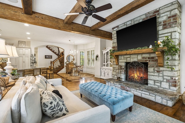 living room with beam ceiling, ceiling fan with notable chandelier, a brick fireplace, wood finished floors, and stairs