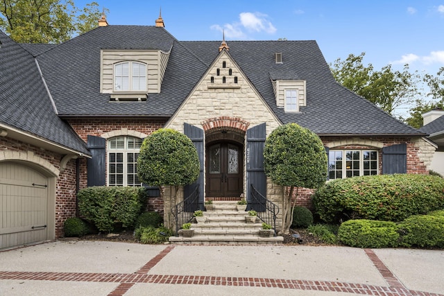 french country inspired facade with driveway, an attached garage, entry steps, brick siding, and roof with shingles