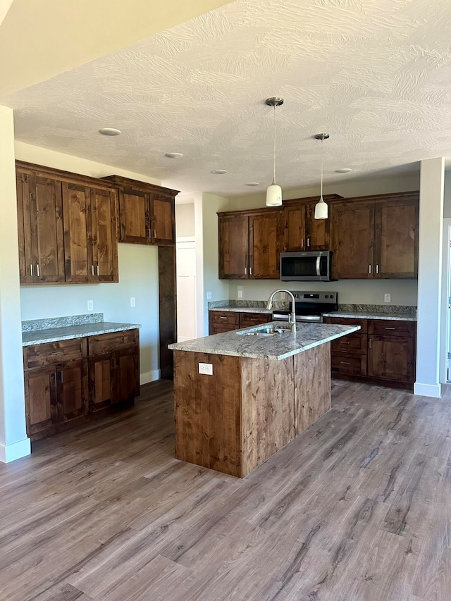 kitchen featuring sink, appliances with stainless steel finishes, hanging light fixtures, light hardwood / wood-style floors, and an island with sink