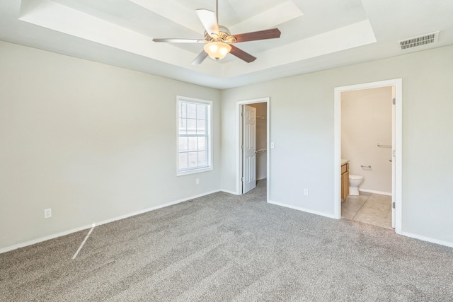 unfurnished bedroom with ceiling fan, ensuite bathroom, a tray ceiling, and a walk in closet