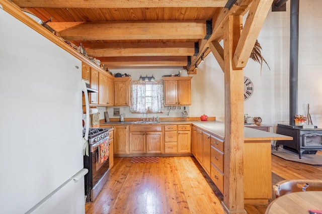 kitchen with white refrigerator, beamed ceiling, light wood-type flooring, stainless steel gas range oven, and a wood stove