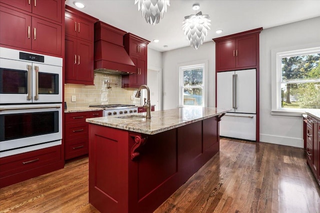 kitchen featuring white appliances, a center island with sink, premium range hood, and a wealth of natural light