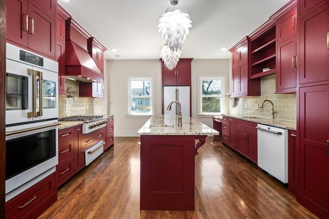 kitchen with an island with sink, stainless steel appliances, premium range hood, dark hardwood / wood-style flooring, and decorative light fixtures