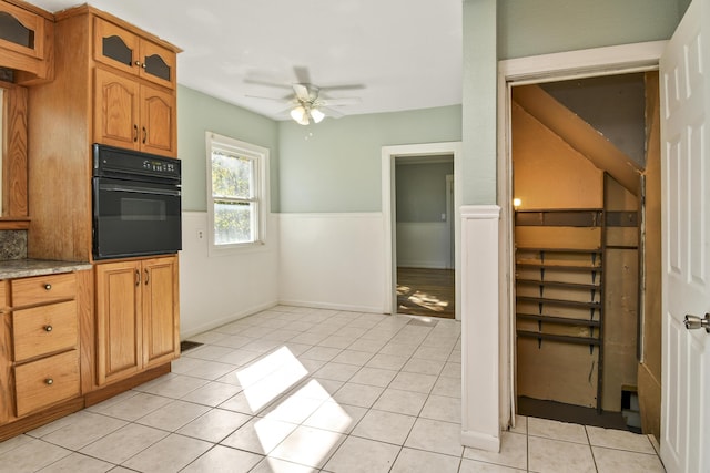 kitchen with light tile patterned floors, ceiling fan, oven, wainscoting, and glass insert cabinets