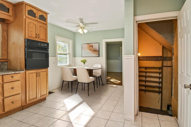 kitchen featuring light tile patterned flooring, a ceiling fan, black oven, wainscoting, and glass insert cabinets