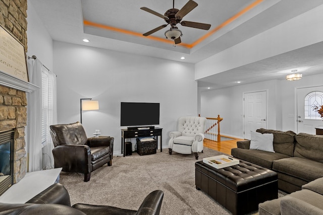 living room featuring ceiling fan, a stone fireplace, a tray ceiling, and carpet