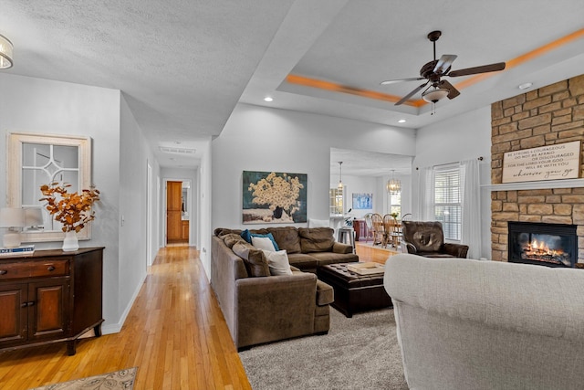 living room featuring ceiling fan, a stone fireplace, a raised ceiling, light hardwood / wood-style flooring, and a textured ceiling