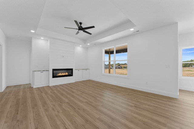 unfurnished living room with light wood-type flooring, a fireplace, ceiling fan, and a tray ceiling