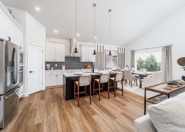 kitchen with wall chimney range hood, stainless steel fridge, a center island with sink, hanging light fixtures, and white cabinetry