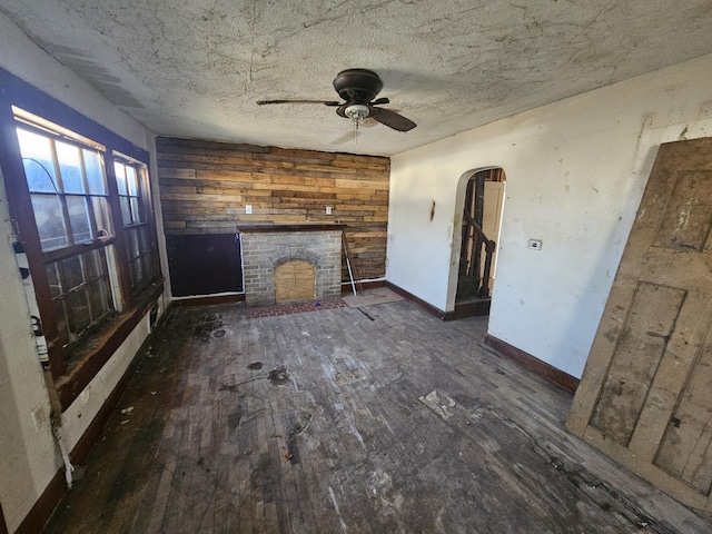 unfurnished living room with arched walkways, a fireplace, ceiling fan, wooden walls, and baseboards