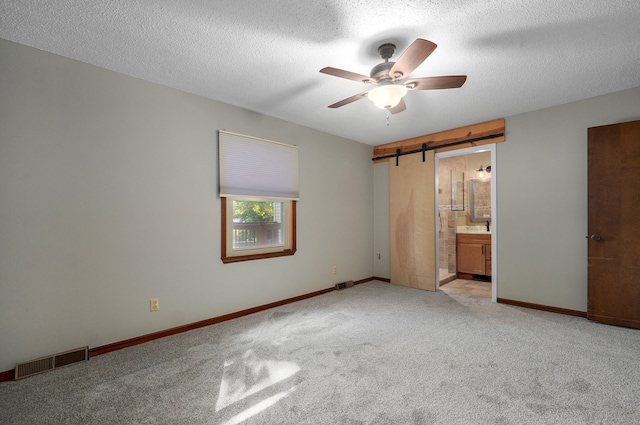 unfurnished bedroom featuring ensuite bathroom, a barn door, a textured ceiling, light carpet, and ceiling fan