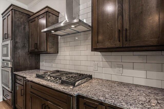 kitchen featuring wall chimney range hood, dark brown cabinets, backsplash, appliances with stainless steel finishes, and light stone counters