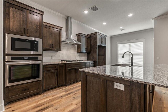 kitchen featuring wall chimney range hood, light hardwood / wood-style flooring, a center island with sink, and crown molding