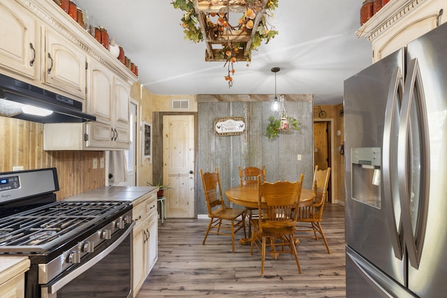 kitchen featuring cream cabinetry, wooden walls, pendant lighting, light wood-type flooring, and appliances with stainless steel finishes