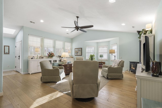 living room with a wood stove, vaulted ceiling with skylight, light hardwood / wood-style floors, and ceiling fan