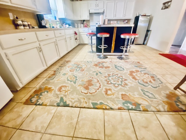 kitchen featuring white cabinets, freestanding refrigerator, light tile patterned floors, a kitchen bar, and under cabinet range hood