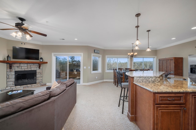 carpeted living room featuring crown molding, a stone fireplace, ceiling fan with notable chandelier, and plenty of natural light