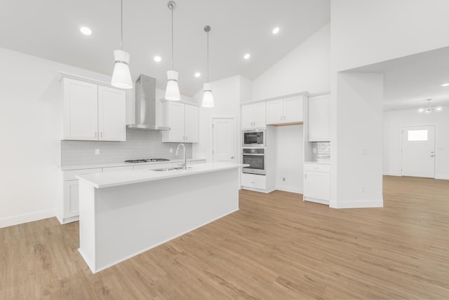 kitchen featuring wall chimney range hood, white cabinets, a center island with sink, appliances with stainless steel finishes, and light wood-type flooring