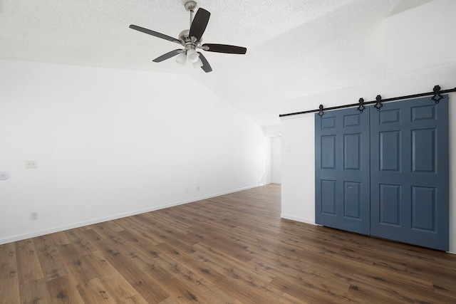 empty room with ceiling fan, a textured ceiling, vaulted ceiling, a barn door, and dark hardwood / wood-style flooring