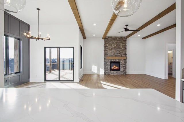 unfurnished living room featuring a stone fireplace, ceiling fan with notable chandelier, beam ceiling, and light hardwood / wood-style flooring