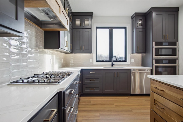 kitchen with sink, custom exhaust hood, light stone counters, light wood-type flooring, and appliances with stainless steel finishes