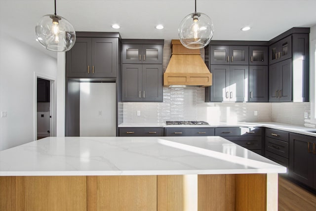 kitchen featuring hanging light fixtures, a kitchen island, custom range hood, stainless steel gas stovetop, and light stone countertops